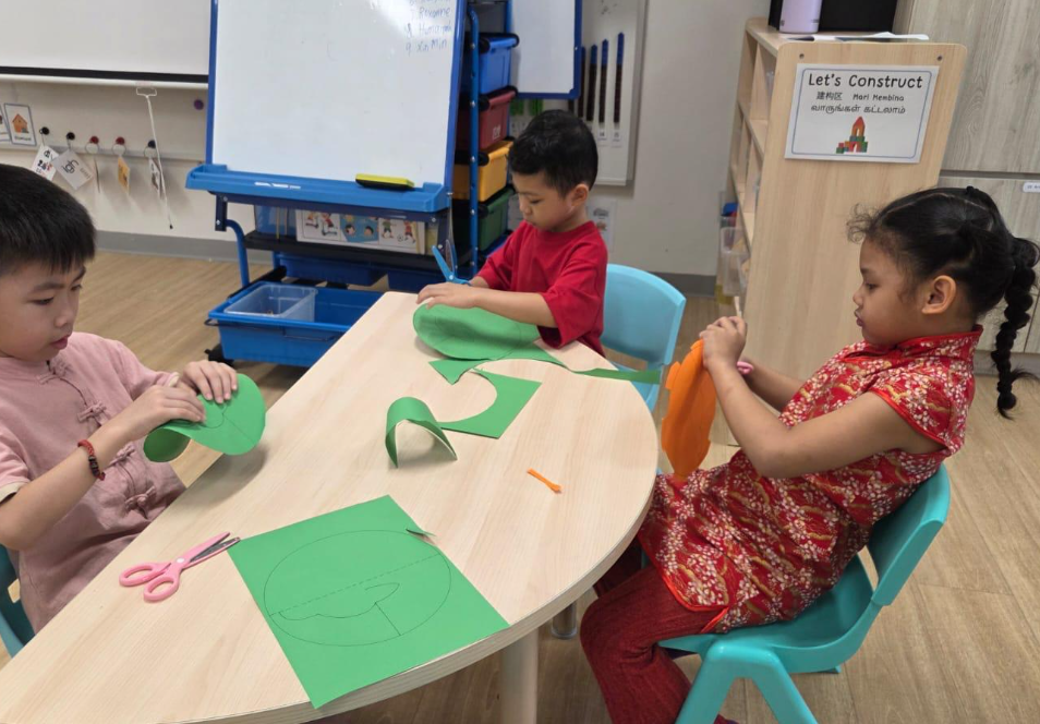 Three children at a classroom table cut green and orange paper shapes with scissors.
